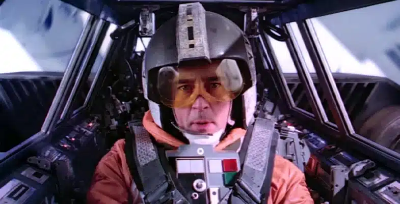 Pilote rebelle en combinaison orange et casque noir avec visière jaune, aux commandes d’un vaisseau dans L’Empire contre-attaque. Il s’agit du personnage Wedge Antilles, incarné par Denis Lawson.