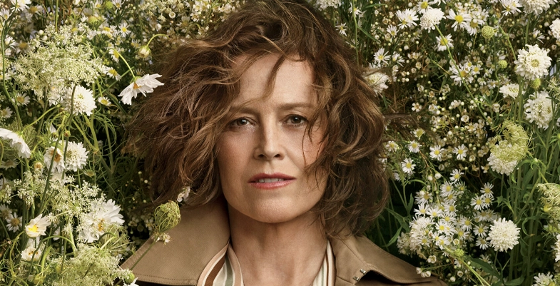 Sigourney Weaver posant parmi des fleurs blanches, avec un regard calme et des cheveux légèrement décoiffés.
