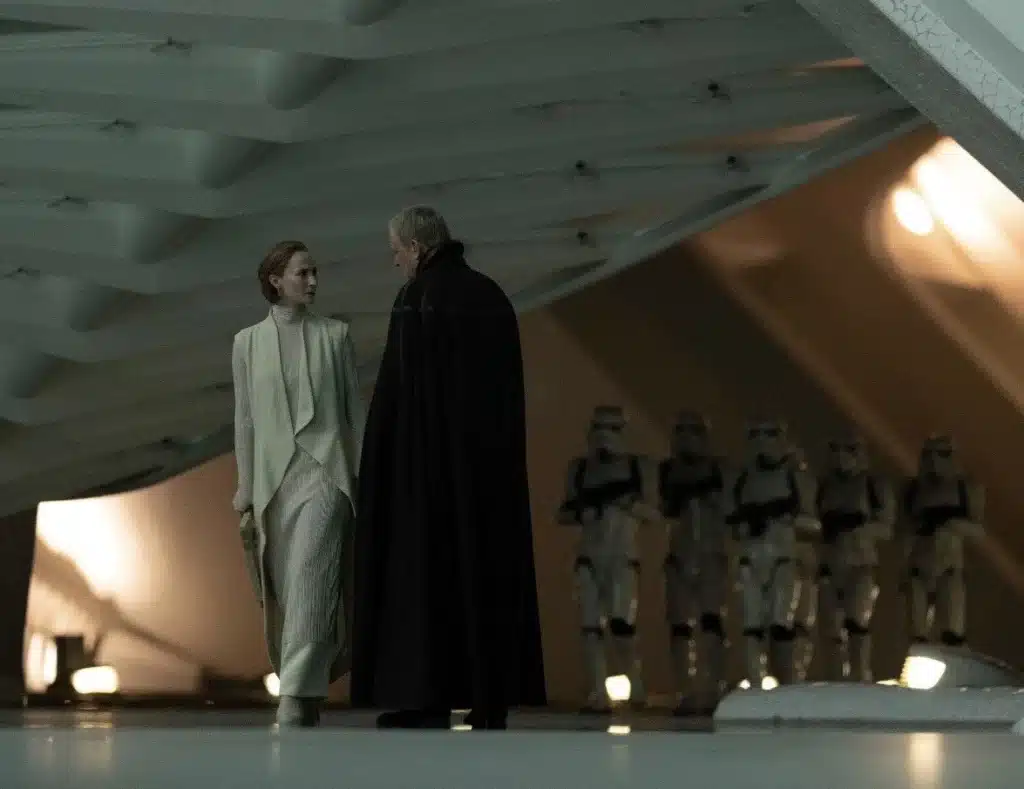 Mon Mothma, vêtue de blanc, discute avec Luthen Rael, portant une cape sombre, sous une structure imposante, avec des stormtroopers en arrière-plan.