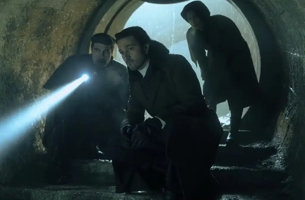 Cassian Andor est au premier plan, éclairé par une torche tenue par un personnage en arrière-plan, dans un tunnel sombre et humide, avec une ambiance mystérieuse et clandestine.
