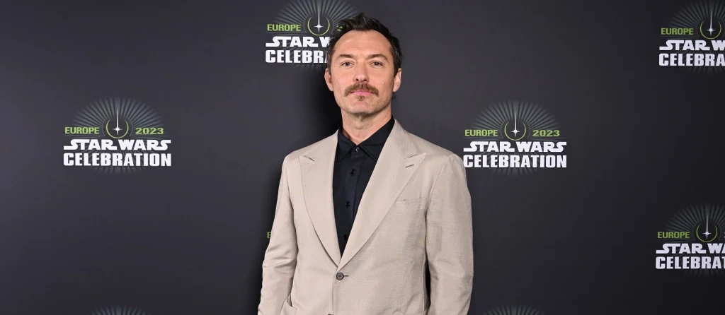 Jude Law vêtu d’un costume beige clair et d’une chemise noire se tient devant un fond promotionnel de Star Wars Celebration Europe.