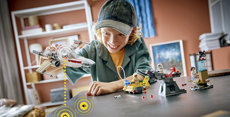 Un adolescent souriant porte une casquette et joue avec des figurines LEGO Star Wars sur une table, en tenant un vaisseau X-Wing. Plusieurs personnages et mini-décors LEGO sont alignés devant lui dans une chambre avec des étagères en arrière-plan.